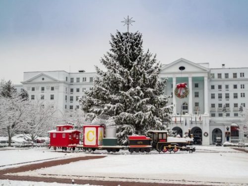 A snowy scene featuring a large Christmas tree, a decorated train, and a festive building in the background.