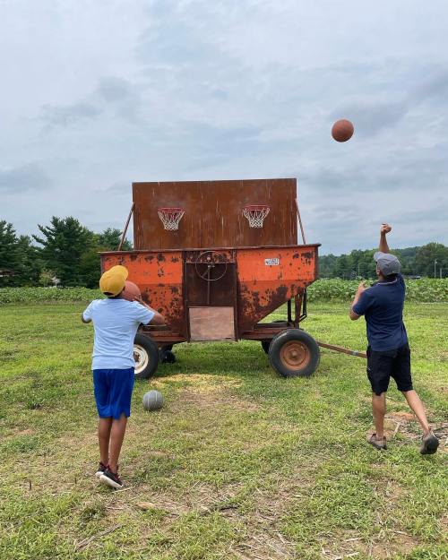Two boys shooting basketballs at a makeshift hoop on a trailer in a grassy field under a cloudy sky.