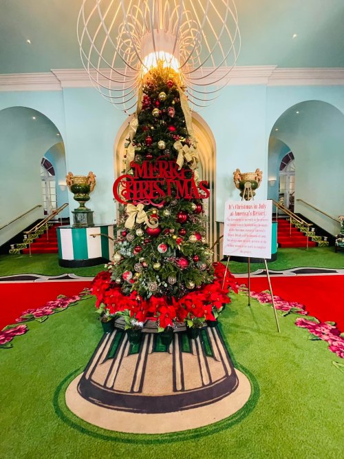 A decorated Christmas tree with a "Merry Christmas" sign, surrounded by red poinsettias in a festive lobby setting.