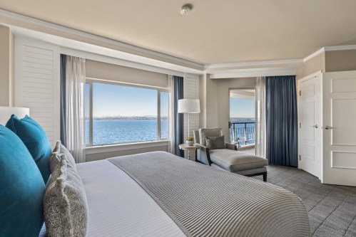 A modern hotel room with a large window overlooking the water, featuring a bed, armchair, and stylish decor.