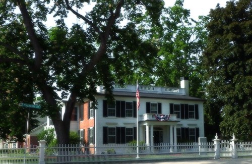 A historic two-story house with a white picket fence, surrounded by trees and featuring an American flag.