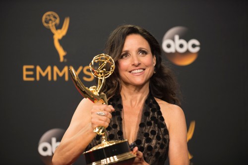 Julia Louis-Dreyfus smiles while holding an Emmy award at the Emmys, wearing a black dress against a backdrop of logos.