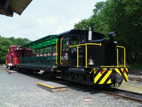 A black and yellow locomotive with a green flatcar parked beside a red train car in a rural setting.