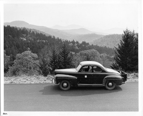 A vintage black car parked on a road with a scenic mountain landscape in the background.