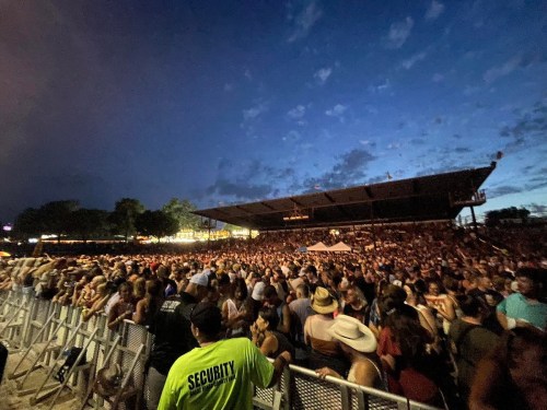 A large crowd gathers at an outdoor concert venue under a twilight sky, with security personnel visible in the foreground.