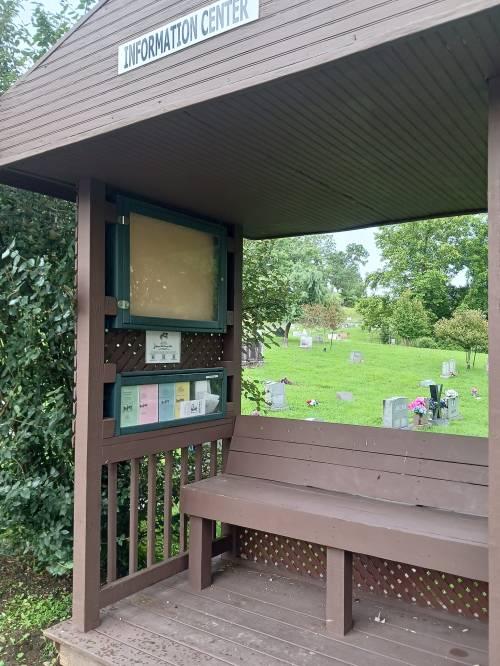 A wooden information center with a bench, surrounded by greenery and gravestones in the background.