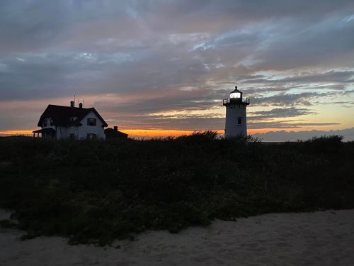 A lighthouse and a house silhouette against a colorful sunset sky, with clouds and grassy foreground.
