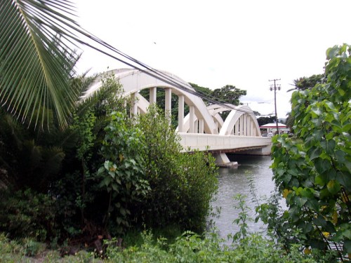 A white arch bridge spans a river, surrounded by lush greenery and trees under a cloudy sky.