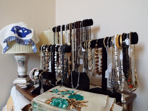 A display of various necklaces and bracelets on black stands, with a lamp and embroidered cloth in the background.