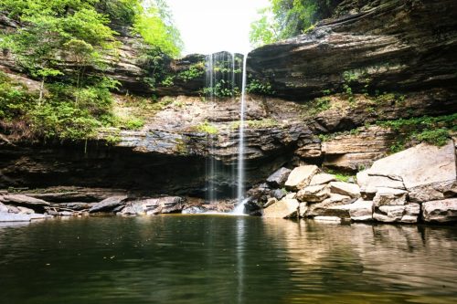A serene waterfall cascades into a calm pool, surrounded by lush greenery and rocky cliffs.