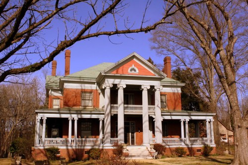 A large, historic brick house with white columns, surrounded by trees and a clear blue sky.