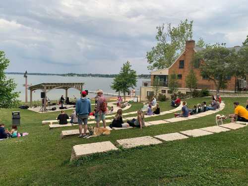 A crowd gathers on a grassy area by a lake, enjoying a performance near a small stage and trees.