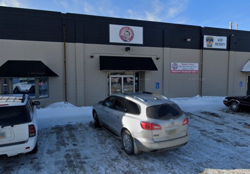 A parking lot with snow, featuring a building with a sign and a few parked cars.