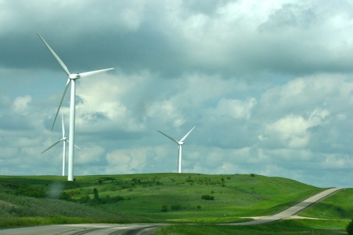 Wind turbines on green hills under a cloudy sky, with a winding road in the foreground.