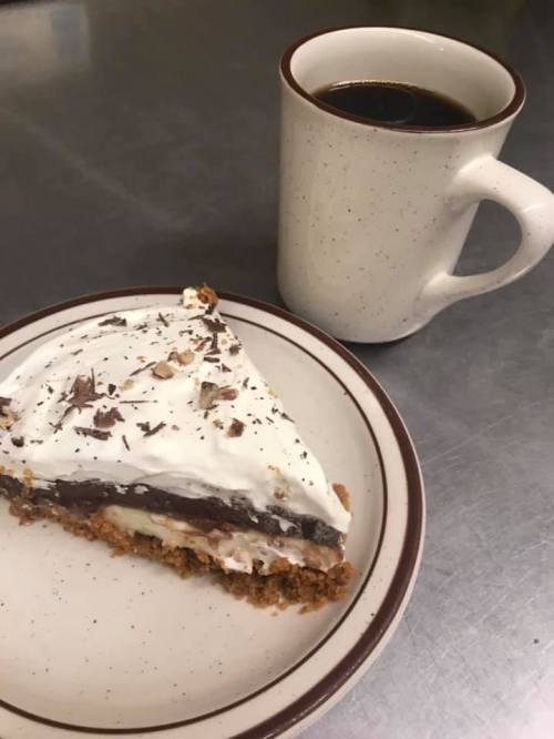 A slice of layered dessert with whipped cream and chocolate shavings, next to a cup of black coffee on a table.