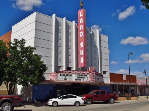 Historic Land Sun theater with a bright marquee, surrounded by trees and parked cars under a blue sky.