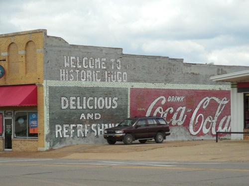 A vintage mural on a building reads "Welcome to Historic Hugo" alongside a Coca-Cola advertisement.