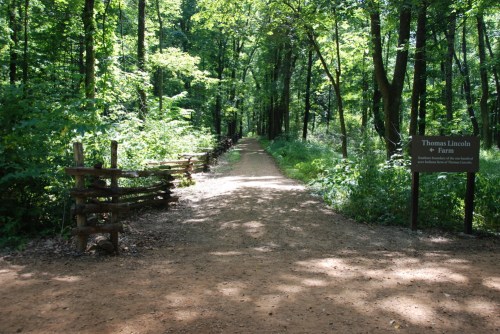 A dirt path through a lush green forest, with wooden fences and a sign for Thomas Lincoln Farm on the right.