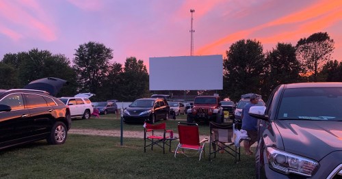 A drive-in movie scene at sunset, with cars parked and a blank screen set up in a grassy area.