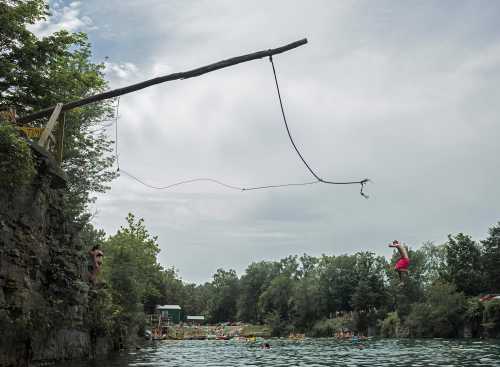 A person jumps off a wooden platform into a lake, with others swimming and relaxing nearby. Trees surround the area.