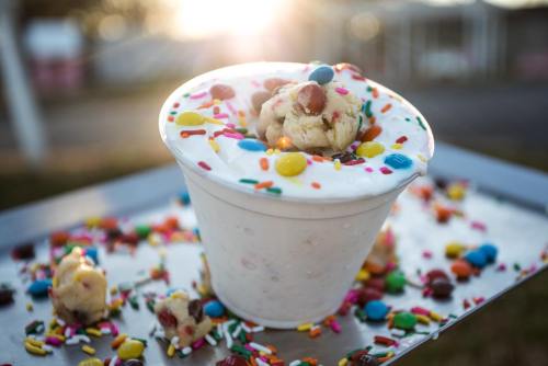 A cup of ice cream topped with colorful sprinkles and cookie dough, surrounded by more sprinkles on a tray.