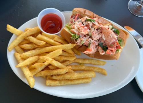 A plate with a lobster roll topped with greens and a side of golden French fries and a small cup of ketchup.