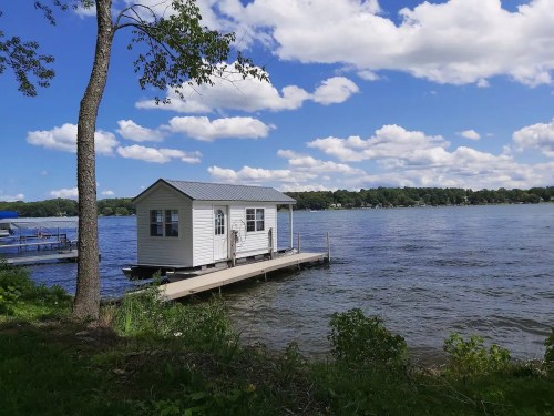 A small white houseboat on a dock by a calm lake, surrounded by trees and fluffy clouds in a blue sky.