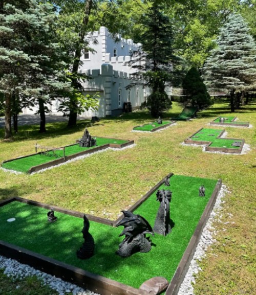 A miniature golf course with themed sculptures, surrounded by green grass and trees, near a castle-like building.
