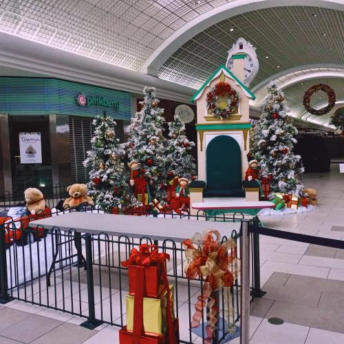 A festive mall scene with decorated Christmas trees, gifts, and a holiday-themed display featuring a small house and teddy bears.