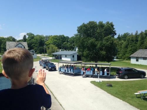 A child waves as a tram filled with people passes by on a sunny day near a park with green trees and buildings.