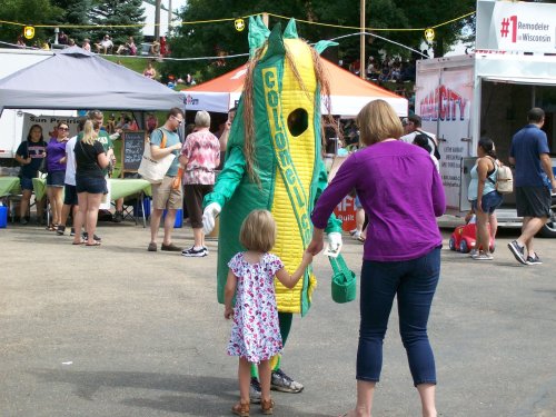 A child in a dress holds hands with a woman as they meet a corn mascot at a lively outdoor event.