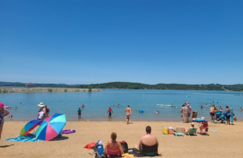 A sunny beach scene with people relaxing on the sand and swimming in the water, surrounded by green hills.