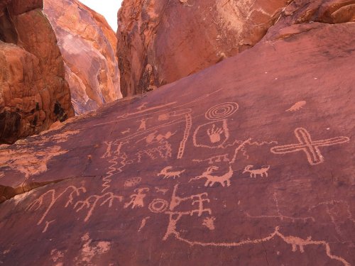 Ancient petroglyphs carved into a red rock surface, depicting figures, animals, and symbols.