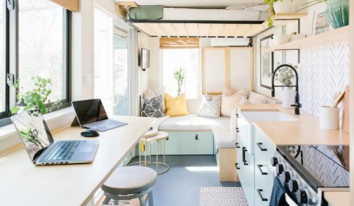 A cozy, modern tiny home interior with a kitchen, workspace, and seating area filled with plants and natural light.