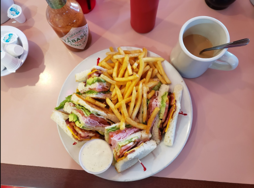 A plate with a club sandwich, fries, a cup of coffee, and a bottle of hot sauce on a pink table.