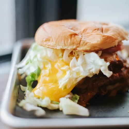 A close-up of a juicy burger with lettuce, tomato, and a runny egg on a soft bun, served on a tray.