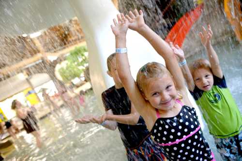 Three children play in a water park, joyfully raising their arms under a waterfall, surrounded by splashing water.