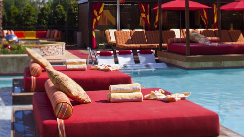 Lounge area by a pool with red cushions, towels, and shaded seating under colorful umbrellas.