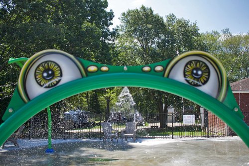 A colorful frog-themed water feature with large eyes, surrounded by a splash pad and trees in the background.