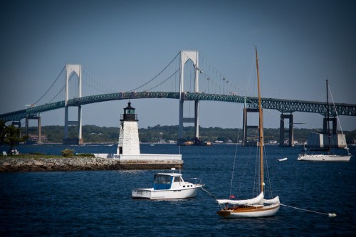 A lighthouse and sailboats in the foreground with a large bridge spanning the water in the background.