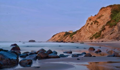 A serene beach scene with smooth rocks, gentle waves, and a cliffside house under a soft blue sky.