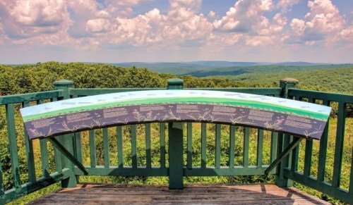A scenic overlook with a curved informational panel, surrounded by lush green hills and a blue sky with clouds.