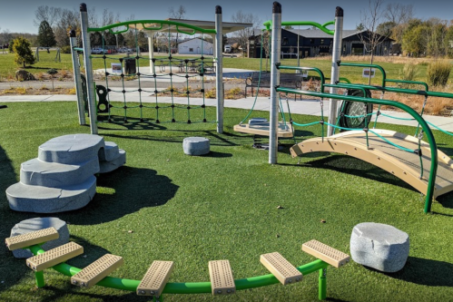 A playground with climbing structures, balance beams, and stepping stones on green turf, surrounded by open space.