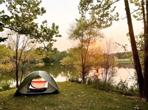 A tent by a serene lake at sunset, surrounded by trees and lush greenery.