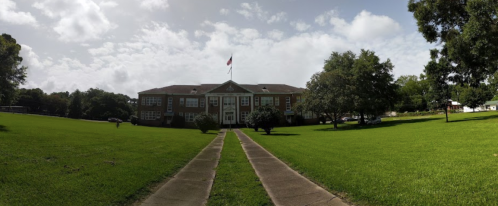 A large brick school building surrounded by green grass and trees, with a pathway leading to the entrance.