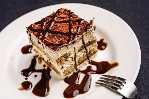 A slice of layered cake drizzled with chocolate sauce on a white plate, with a fork beside it.