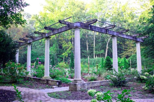 A serene garden with a stone pergola supported by columns, surrounded by lush greenery and blooming flowers.