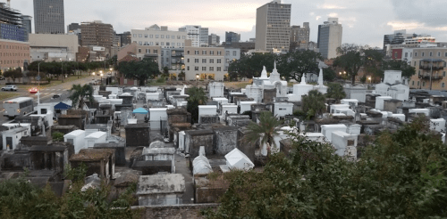 Aerial view of a cemetery with white tombs, surrounded by city buildings and greenery at dusk.