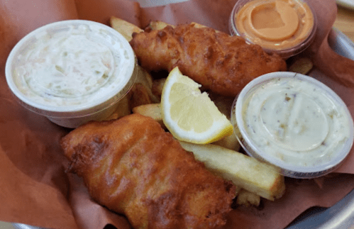 A basket of golden fried fish fillets and fries, served with two dipping sauces and a lemon wedge.