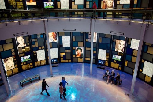 A modern exhibition space with colorful screens displaying portraits and text, featuring visitors walking on a reflective floor.
